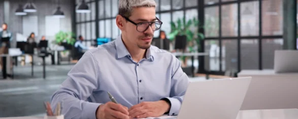 Man with glasses writing notes at a desk while reviewing content on a laptop in a modern office