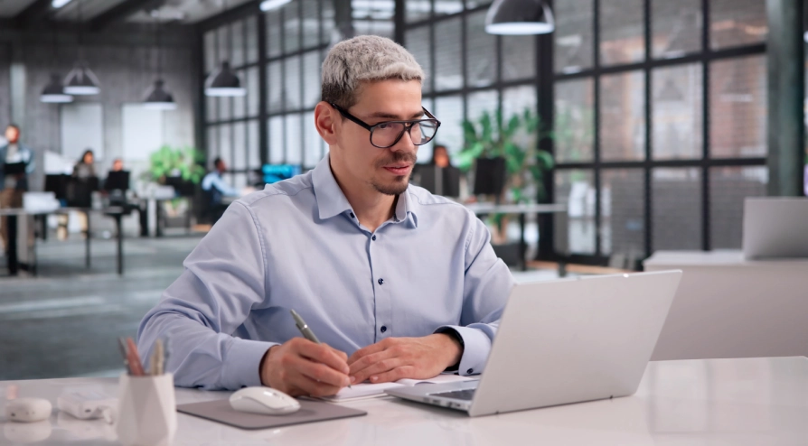 Man with glasses writing notes at a desk while reviewing content on a laptop in a modern office