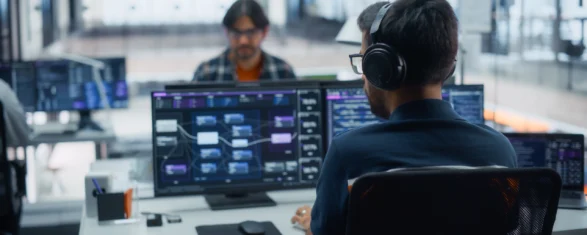 Engineer wearing headphones working at a workstation with workflow dashboards on two monitors in an open-plan office